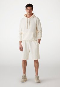 Cremefarvet hoodie med et tekstureret mønster, frontlommer og snore; parret med matchende shorts. Båret med lyse sneakers.