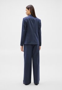 Žena stojící zády, oblečená v modrém šitém saku a širokých kalhotách na světlém jednobarevném pozadí.