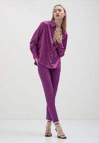 Blusa di raso fucsia con frontale a bottoni e maniche lunghe, abbinata a pantaloni slim-fit coordinati e tacchi neri con cinturini.