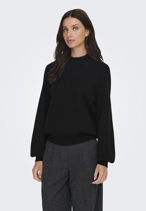 Donna con capelli lunghi e scuri, indossa un maglione nero a maniche lunghe e pantaloni grigi a vita alta e gamba larga, in piedi davanti a uno sfondo semplice.