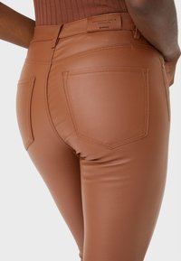 Braune Kunstlederhose mit glatter Textur, die zwei Gesäßtaschen und sichtbare Nähte aufweist. Das Etikett trägt die Aufschrift "Authentic Denim Supplier".