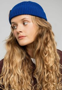 Niebieska wełniana czapka z teksturalnym wykończeniem i małą skórzaną metką, noszona przez osobę z długimi, falującymi blond włosami.