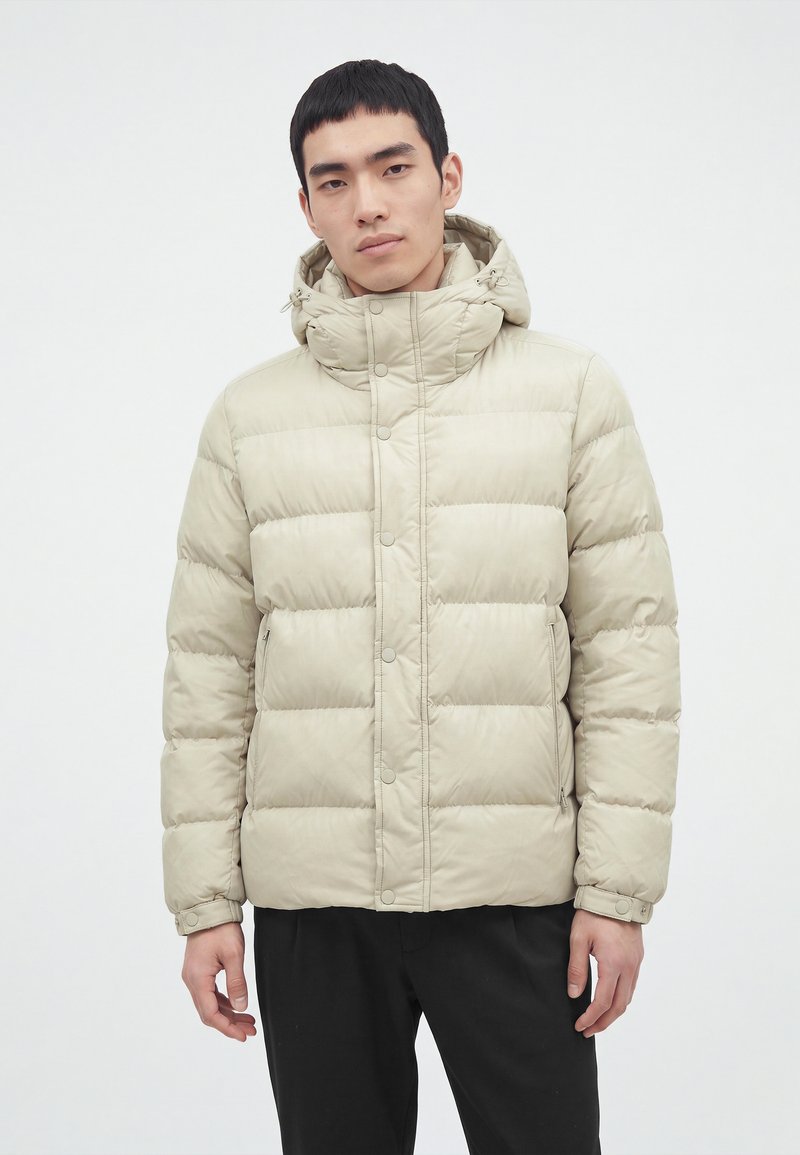 Jeune homme portant une doudoune beige avec capuche et un pantalon noir, debout devant un fond blanc uni.