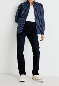 Person klädd i en mörkblå randig skjorta över vit t-shirt, svarta byxor och vita låga sneakers som står mot en vit vägg.