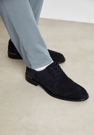 Schwarze Schnür-Damenschuhe aus Wildleder, getragen mit hellblauen Hosen und weißen Socken auf einem hellbeigen Boden.