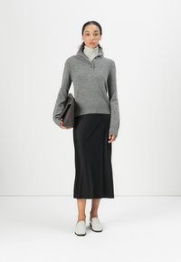 Pull en laine gris avec un col replié et des boutons, jupe midi noire, chaussures gris à enfiler, tenant une pochette marron. Textures lisses dans l'ensemble.