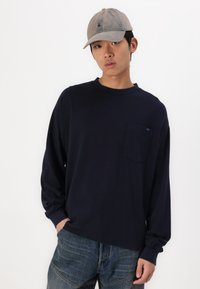 Μπλέ μακρυμάνικο μπλουζάκι με τσέπη στο στήθος, συνδυασμένο με ξεθωριασμένα τζιν και γκρι καπέλο μπέιζμπολ με λογότυπο.