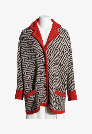Cardigan in maglia con motivo geometrico nero e bianco, colletto, polsini e tasche rossi, chiusura con bottoni, esposto su un manichino bianco.