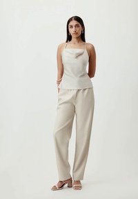 Žena stoji u bež topu bez rukava s tankim naramenicama, odgovarajućim širokim hlačama visokog struka i bež sandalama na petu, na jednobojnoj pozadini.