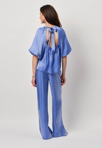 Blauwe satijnen top met een rugkeyhole en strikdetail, gecombineerd met bijpassende losse broek, beide met een soepele, glanzende textuur.