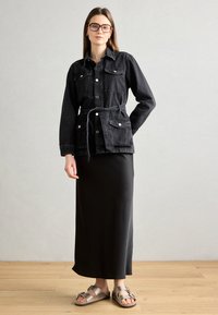 Jachetă din denim negru cu buzunare frontale, talie cu curea și accesorii din argint, purtată cu o fustă neagră elegantă și sandale maro.