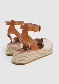 Beige kanvoso espadrilės su rudais odiniais kulkšnies dirželiais, austa jūrinėmis padais ir šiek tiek pakeltu kulnu patogumui. Minimalistinis dizainas.