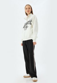 Sudadera blanca con texto gráfico negro, combinada con pantalones de chándal negros con rayas laterales rosas y zapatillas blancas con suelas beige.