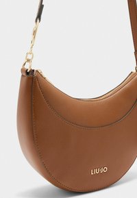 Bolso bandolera de cuero marrón con forma redondeada, herrajes dorados y cierre de cremallera. Presenta costuras sutiles y un logo de marca.