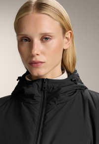 Zwarte capuchonjas met een ritssluiting, zachte textuur en verstelbare trekkoorden bij de halslijn, geplaatst tegen een neutrale achtergrond.