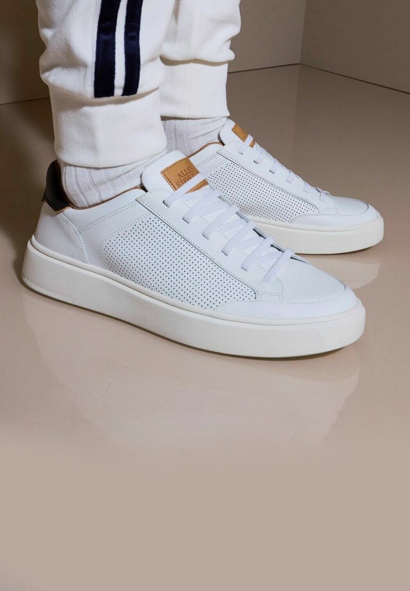 Weiße Ledersneaker mit durchlässigen seitlichen Einsätzen, flachen Schnürsenkeln und einem braunen Lederakzent an der Ferse. Dicke weiße Gummisohlen und ein minimalistisches Design.