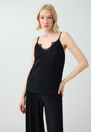 Frau mit schulterlangem blondem Haar trägt ein schwarzes plissiertes Camisole mit Spitzenbesatz und dazu passende plissierte Hose, stehend vor hellem Hintergrund.