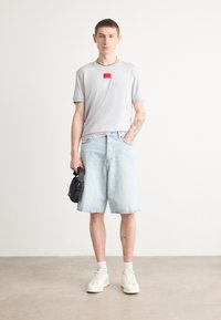 T-shirt grigio a maniche corte con un'etichetta logo rossa, abbinato a pantaloni corti in denim azzurri. Gli accessori includono una borsa nera e sneakers bianche.
