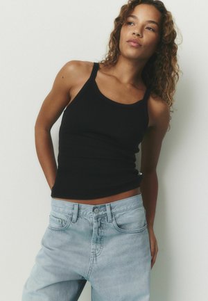 Junge Frau mit lockigem Haar, die ein schwarzes Tanktop und hellblaue Jeans trägt, vor einem schlichten weißen Hintergrund stehend.