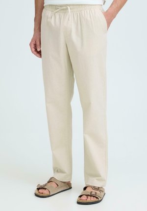 Pantalon beige taille élastique porté avec des sandales beige à bout ouvert, modèle debout avec une main dans la poche sur un fond uni.
