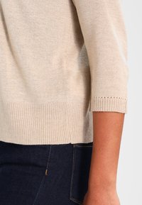 Gebreide trui in lichtbeige met een geribbelde zoom en manchetten, gekenmerkt door een zachte textuur en mouwen tot aan de ellebogen. Gecombineerd met donkere jeans.