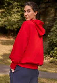 Sudadera de algodón roja con un bolsillo frontal y capucha con cordón, combinada con pantalones azul marino, con un corte relajado y una textura suave frente a un fondo natural.