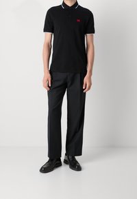 Svart pikétröja med vit kant och liten röd logotyp, ihop med lösa svarta byxor. Outfiten har klassiska, minimalistiska designelement.