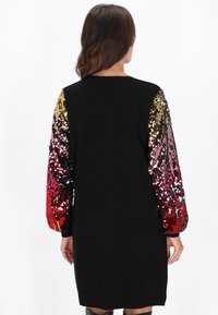 Czarna sukienka z długimi rękawami, ozdobiona wielokolorowymi cekinami, przechodzącymi od złota, przez róż do czerwieni. Gładka tekstura i luźny krój.
