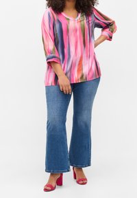 Blouse multicolore avec des rayures verticales en rose, bleu, vert et orange ; col en V, manches trois-quarts, portée avec un jean évasé et des talons roses.