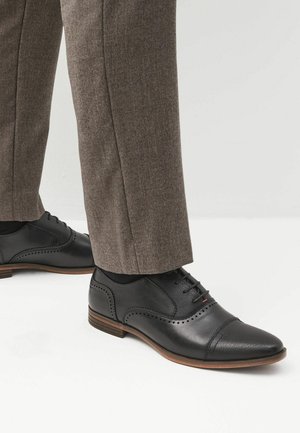 Zapatos de vestir de cuero negro con punta afilada, perforaciones decorativas tipo brogue, y una suela de madera marrón contrastante, combinados con pantalones marrones.