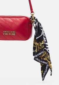 Borsa in pelle trapuntata rossa con logo dorato, dotata di una sciarpa staccabile nera e dorata a motivo legato alla tracolla. Design compatto e rettangolare.