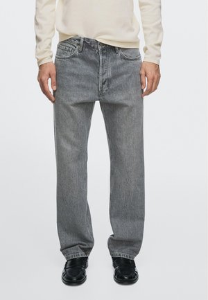 Man met lichtbeige shirt met lange mouwen, losse grijze spijkerbroek en zwarte leren schoenen, staand tegen een effen witte achtergrond.