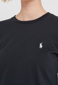 Musta puuvillainen t-paita, jossa on pyöreä kaula-aukko ja lyhyet hihat. Vasemmassa rinnassa on pieni valkoinen kirjailtu logo, jossa on ratsastaja.