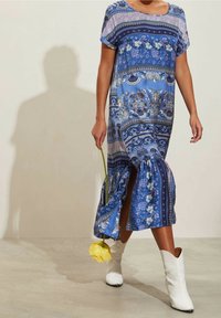 Modré květinové maxi šaty s krátkými rukávy, bočním rozparkem a volánkovým zakončením. Doplněné bílými kovbojskými botami a žlutou květinou.