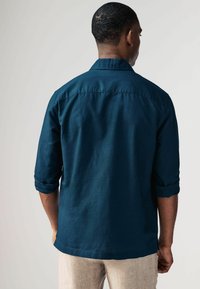 Camisa de botones de manga larga en un tono verde azulado oscuro, con mangas remangadas y dobladillo recto. Textura suave con un ligero brillo y un diseño de corte relajado.