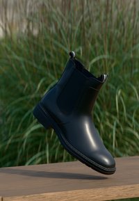 Botte Chelsea en cuir noir suspendue au-dessus d'une surface en bois avec de l'herbe verte floue en arrière-plan.
