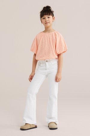 Jeune fille debout, mains détendues, portant un haut pêche à manches bouffantes, un pantalon blanc évasé et des chaussures beige à enfiler, sur un fond beige.