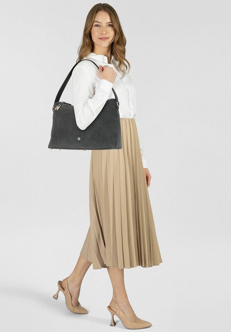 Borsa in pelle scamosciata grigia con design strutturato, ferrature argentate e tracolla lunga, tenuta da una persona con camicia bianca e gonna beige plissettata.