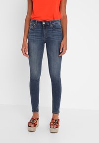 Vaqueros skinny de mezclilla en azul oscuro con un ligero desvanecimiento, de cintura alta y diseño de cinco bolsillos, combinados con sandalias de plataforma florales.