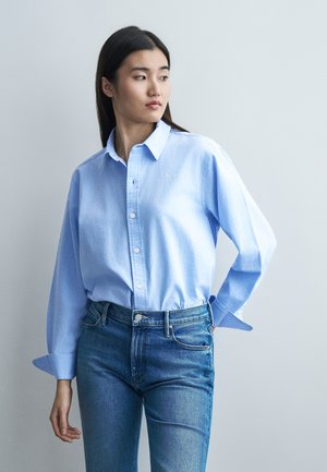 Νεαρή γυναίκα με μακριά σκούρα μαλλιά, φορώντας ένα ανοιχτό μπλε πουκάμισο κουμπωμένο μέσα σε ψηλόμεσο μπλε τζιν παντελόνι, κοιτάζοντας προς το πλάι.