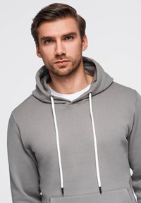 Šedá mikina z měkkého materiálu s kapucí na šňůrku a bílými akcenty. Obsahuje klokaní kapsu a klasický střih.