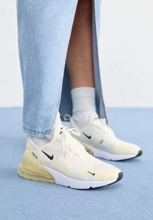 Personne portant des baskets Nike Air Max 270 jaune clair avec des chaussettes blanches et un jean large bleu clair sur un sol bleu.