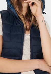 Námořnický modrý péřový vest, který má kapuci, přední zip, prošívanou texturu a kontrastní vnitřní podšívku, nošený přes světlý top.