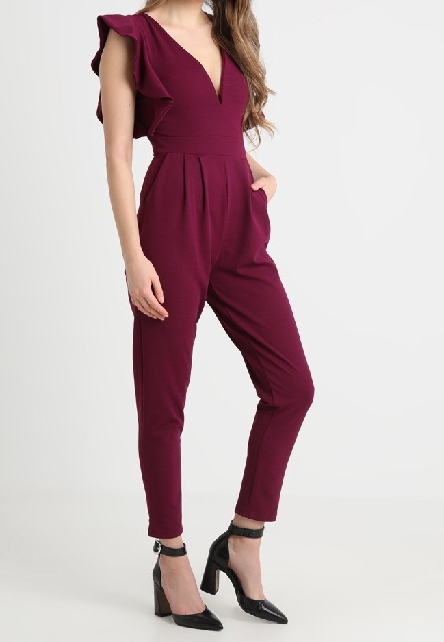 Femme portant une combinaison bordeaux sans manches avec épaules à volants et des talons noirs à bride à la cheville, debout devant un fond clair uni.