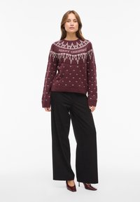 Maroon knit sweater with white “MERRY CHRISTMAS” text, geometric patterns, and long sleeves, paired with black wide-leg trousers.