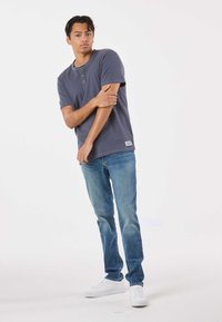 Maglietta henley da uomo color grigio scuro con maniche corte, dotata di due bottoni in legno, abbinata a jeans azzurri e sneakers bianche.