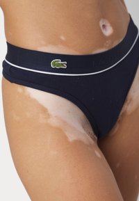 Navy blue bikini bottoms with a ribbed texture, featuring an elastic waistband with a white trim and a green Lacoste crocodile logo.