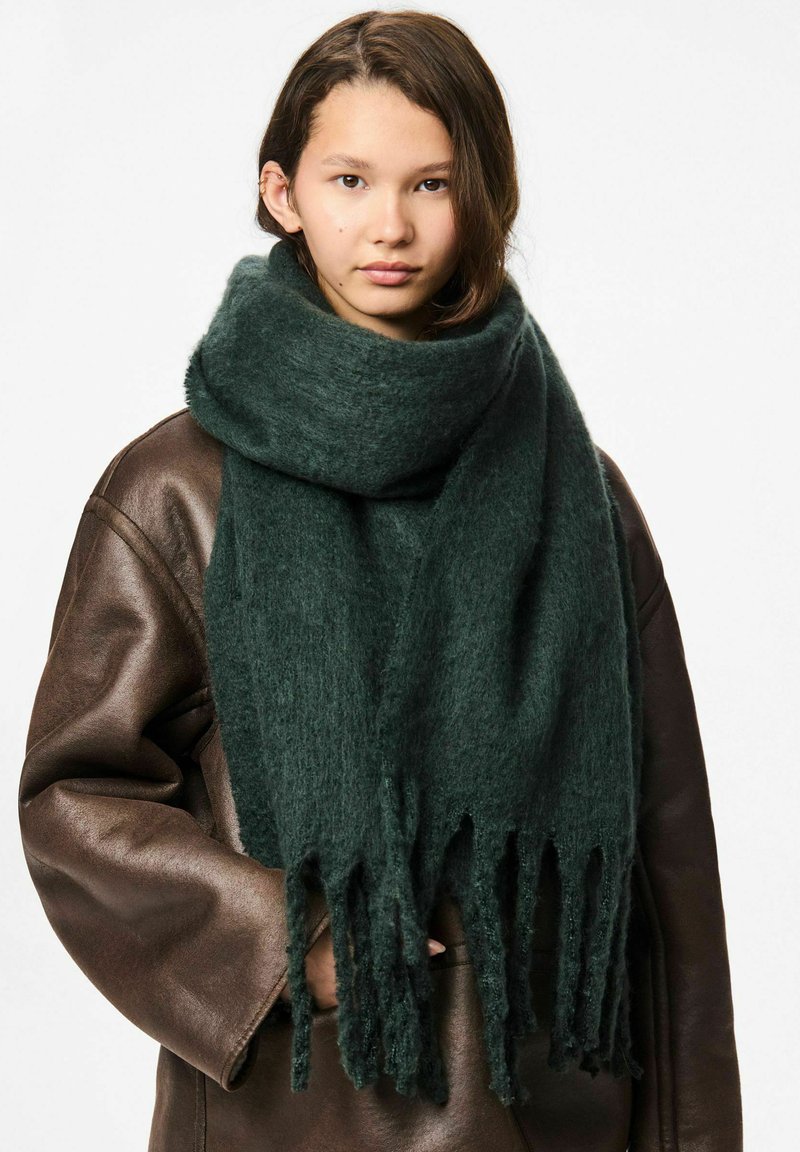 Scarf verde scuro realizzato in tessuto testurizzato, con estremità sfrangiate. Indossato sopra una giacca di pelle marrone, si adagia dolcemente attorno al collo.