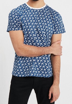 Blå T-shirt med korte ærmer, der har et gentaget mønster af hvide palmer. Rund hals og glat tekstur, båret med sorte bukser.