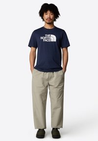 Navyblaues T-Shirt mit weißem Logo, kombiniert mit hellbeigen, lockeren Hosen. Das Outfit wird mit schwarzem Schuhwerk abgerundet.